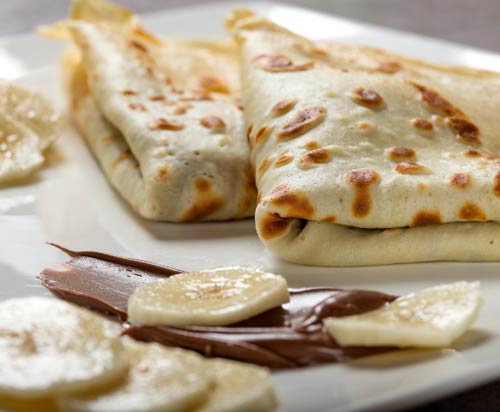 Zwei gefaltete Crêpes auf einem weißen Teller mit Bananenscheiben und einem Aufstrich aus Schokoladen-Haselnuss-Sauce - perfekt für jede Veranstaltung oder als Teil von Reicherts mobiles Catering.