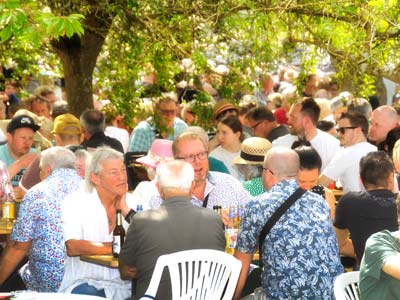 Eine große Gruppe von Menschen sitzt an Tischen im Freien unter Laubbäumen und genießt Essen und Getränke bei einer lebhaften Maifest-Veranstaltung. Die Szene ist voller Gespräche und entspannter Geselligkeit an einem sonnigen Tag.