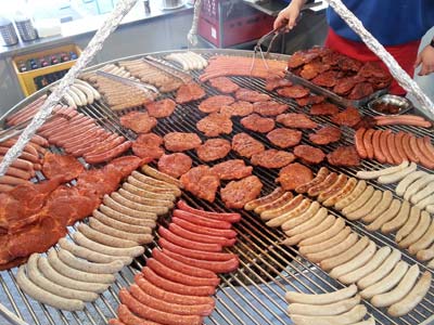 Bei einer lebhaften Maifest-Veranstaltung brutzelt ein großer runder Grill mit Würsten und mariniertem Fleisch, während eine Person mit einer Zange das Essen wendet und so den festlichen Geist der Reicherts-Versammlungen einfängt.